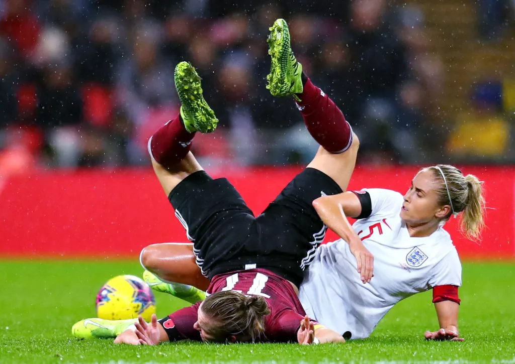 Contrasto duro tra due calciatrici a terra sotto la pioggia: una in maglia scura con i tacchetti verso l'alto, l'altra in bianco con il numero 5 sulla schiena, il pallone giallo sul lato sinistro del campo. - © Catherine Ivill - Getty Images