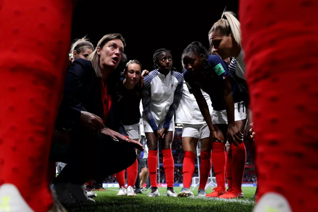Una staff tecnica accovacciata parla alle calciatrici della nazionale francese prima del calcio d'inizio, inquadrata dal basso tra i montanti rossi della porta, il campo illuminato sullo sfondo.