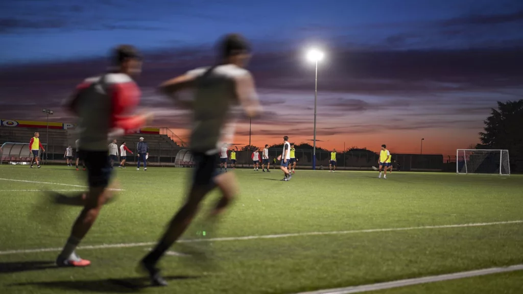Due giocatori in corsa resi mossi dal movimento, cielo arancione del tramonto e riflettore acceso sullo sfondo.
Michele Calvanese — Rangers Qualiano — SPORTSHOTS Magazine