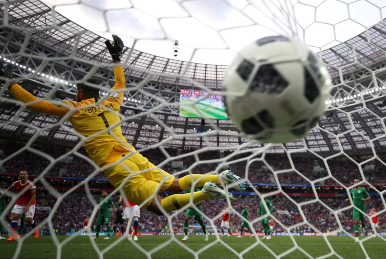 Un portiere in maglia gialla con la scritta "Abdullah" si tuffa inutilmente mentre il pallone entra in rete, fotografato dall'interno della porta con la rete in primo piano e lo stadio sullo sfondo.