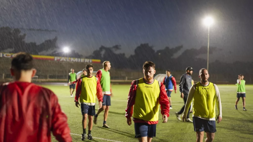 Giocatori con pettorine gialle e rosse camminano su un campo in erba sintetica illuminato dai riflettori, cielo scuro sullo sfondo. Michele Calvanese — Rangers Qualiano — SPORTSHOTS Magazine