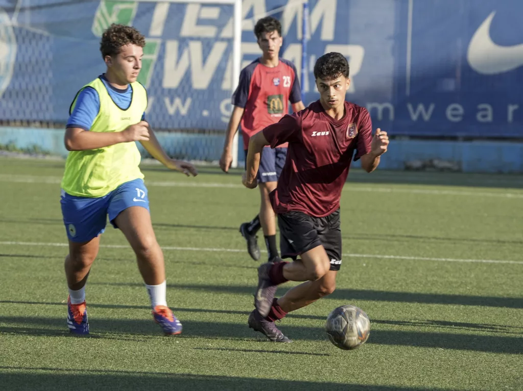 Due giocatori, uno in pettorina gialla e uno in maglia bordeaux, si contendono il pallone su un campo sintetico. Michele Calvanese — Rangers Qualiano — SPORTSHOTS Magazine