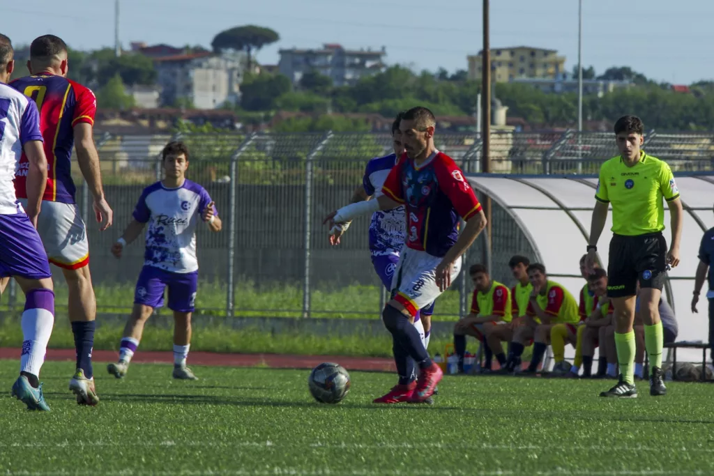 Calciatori in maglia bianca e viola e in maglia rossoblù in azione, arbitro in verde e panchina dietro la recinzione. Michele Calvanese — Rangers Qualiano — SPORTSHOTS Magazine