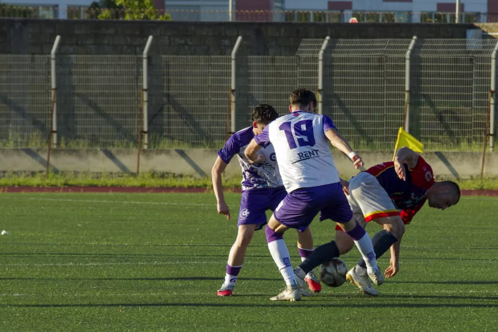 Giocatore con il numero 19 in maglia bianca e viola a contatto con un avversario in maglia rossa che perde l'equilibrio. Michele Calvanese — Rangers Qualiano — SPORTSHOTS Magazine