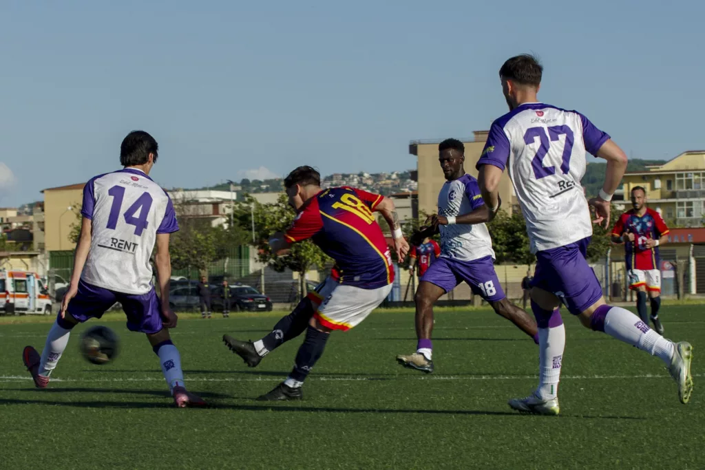 Tre calciatori in corsa su un campo sintetico sotto un cielo azzurro, case sullo sfondo. Michele Calvanese — Rangers Qualiano — SPORTSHOTS Magazine