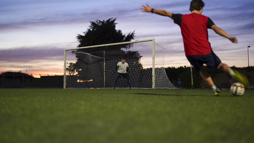 Giocatore in maglia rossa colpisce il pallone in controluce, porta e alberi sullo sfondo al tramonto. Michele Calvanese — Rangers Qualiano — SPORTSHOTS Magazine