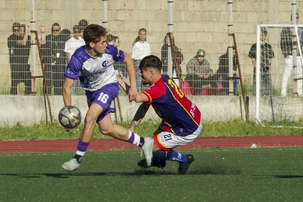 Giocatore in maglia bianca e viola contrasta un avversario in maglia rossoblù che cade, pubblico sullo sfondo. Michele Calvanese — Rangers Qualiano — SPORTSHOTS Magazine