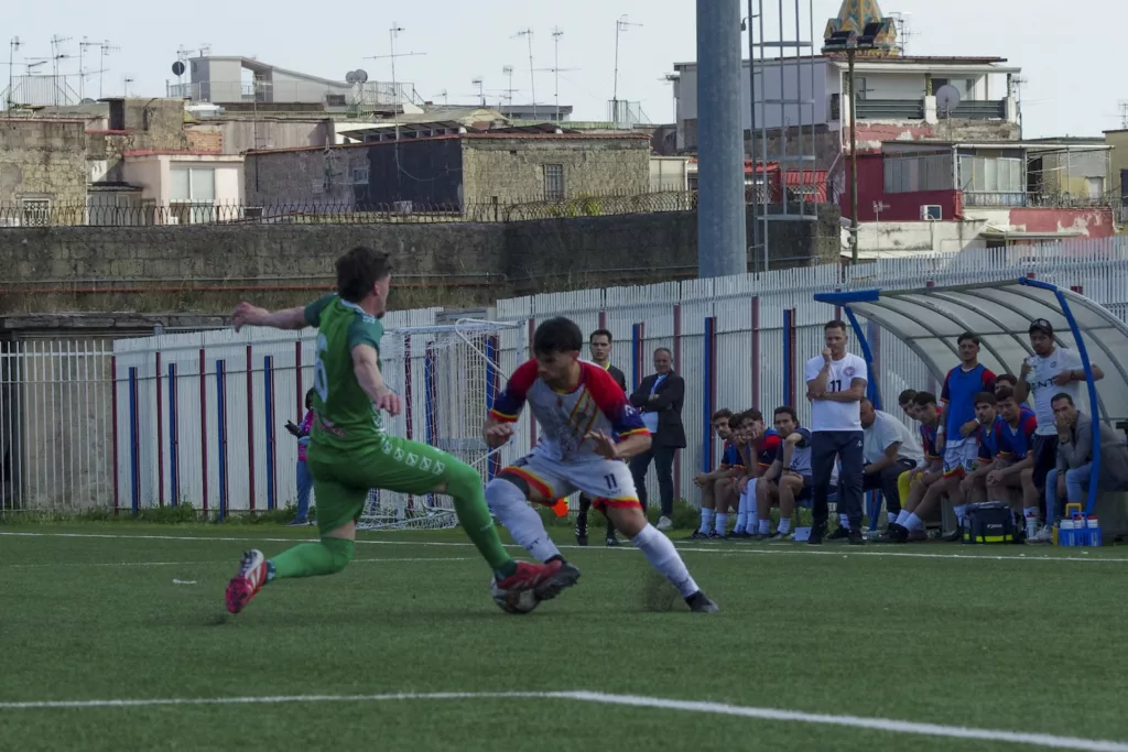 Giocatore in maglia verde e giocatore in maglia bianca in scivolata sul campo sintetico, panchine e palazzine sullo sfondo. Michele Calvanese — Rangers Qualiano — SPORTSHOTS Magazine