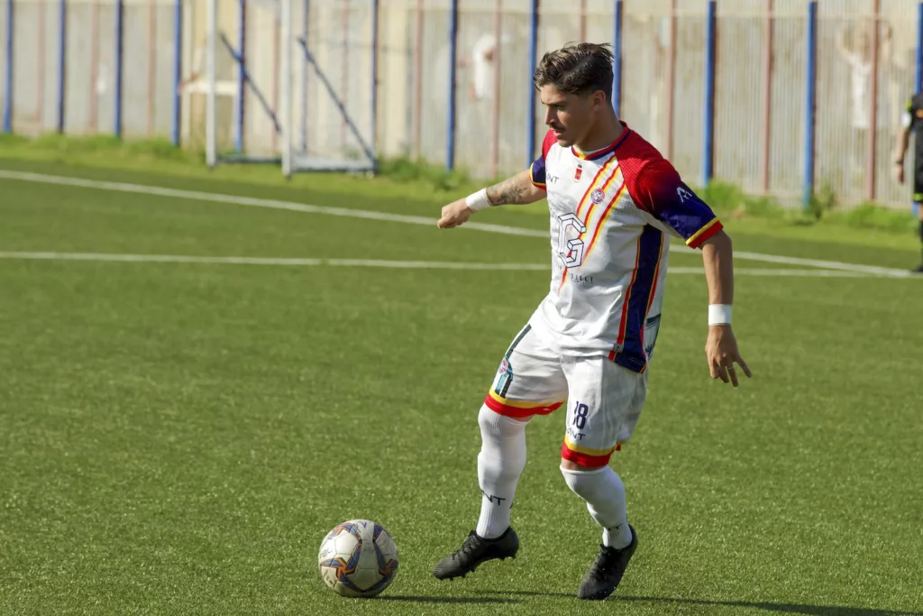 Calciatore in maglia bianca con dettagli rossi e gialli conduce il pallone su un campo in erba. Michele Calvanese — Rangers Qualiano — SPORTSHOTS Magazine