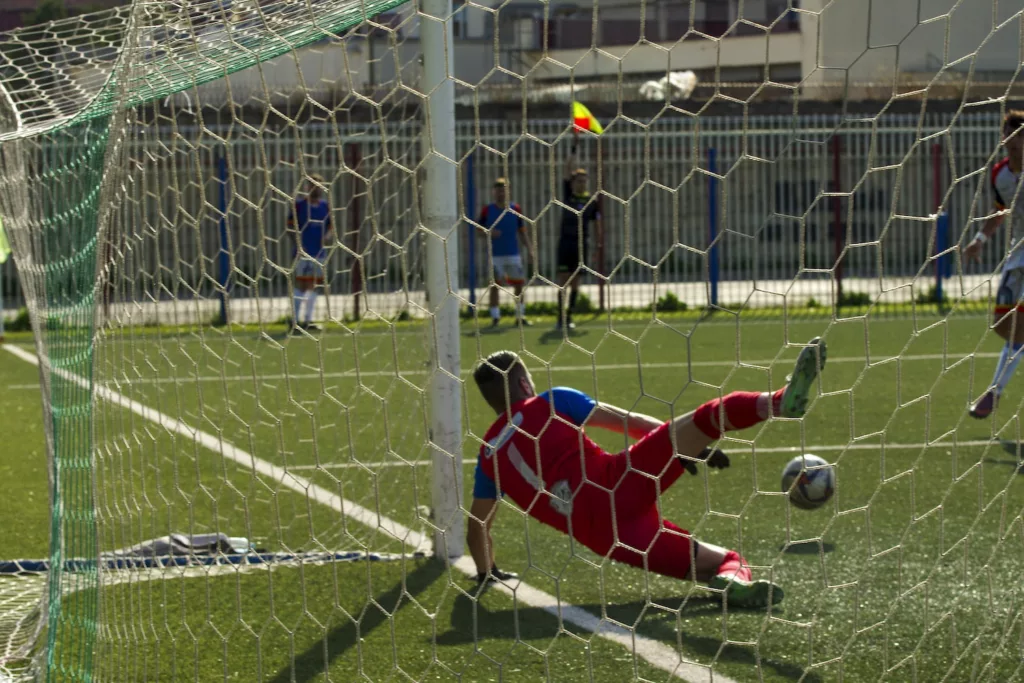 Portiere in maglia rossa in tuffo sul terreno di gioco, visto attraverso la rete della porta. Michele Calvanese — Rangers Qualiano — SPORTSHOTS Magazine