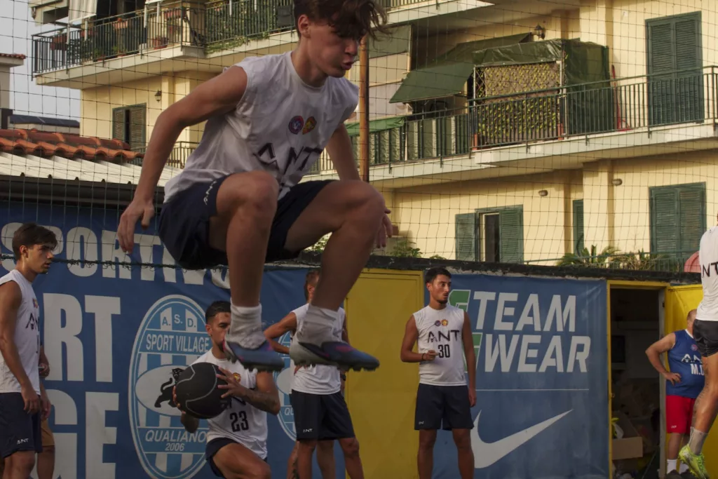 Giocatore in maglia bianca salta sopra un compagno, grande striscione pubblicitario e palazzine sullo sfondo. Michele Calvanese — Rangers Qualiano — SPORTSHOTS Magazine