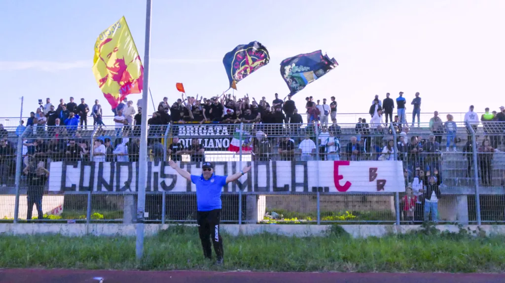 Tifosi in tribuna con bandiere e striscioni, cielo azzurro sullo sfondo. Michele Calvanese — Rangers Qualiano — SPORTSHOTS Magazine
