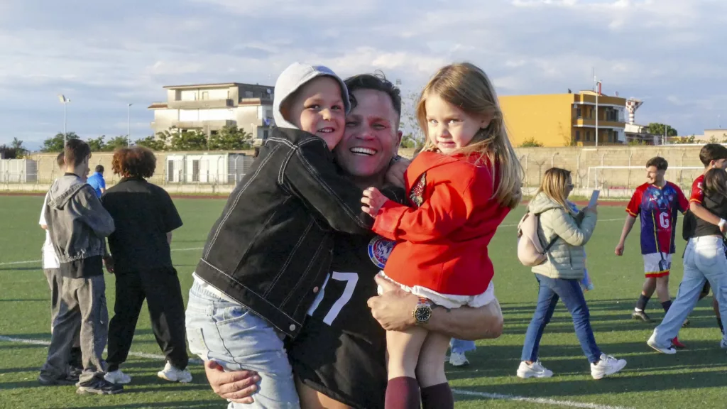 Un uomo e una donna abbracciano una bambina vestita di rosso sul terreno di gioco, altre persone intorno. Michele Calvanese — Rangers Qualiano — SPORTSHOTS Magazine