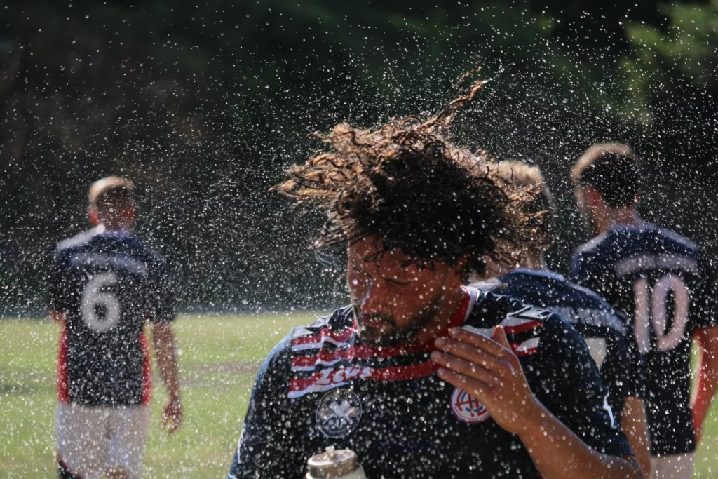 Giocatore con i capelli lunghi bagnati e spruzzi d'acqua, compagni in maglia scura sullo sfondo. Sofia Gasbarri — ASD Montorio Romano — SPORTSHOTS Magazine