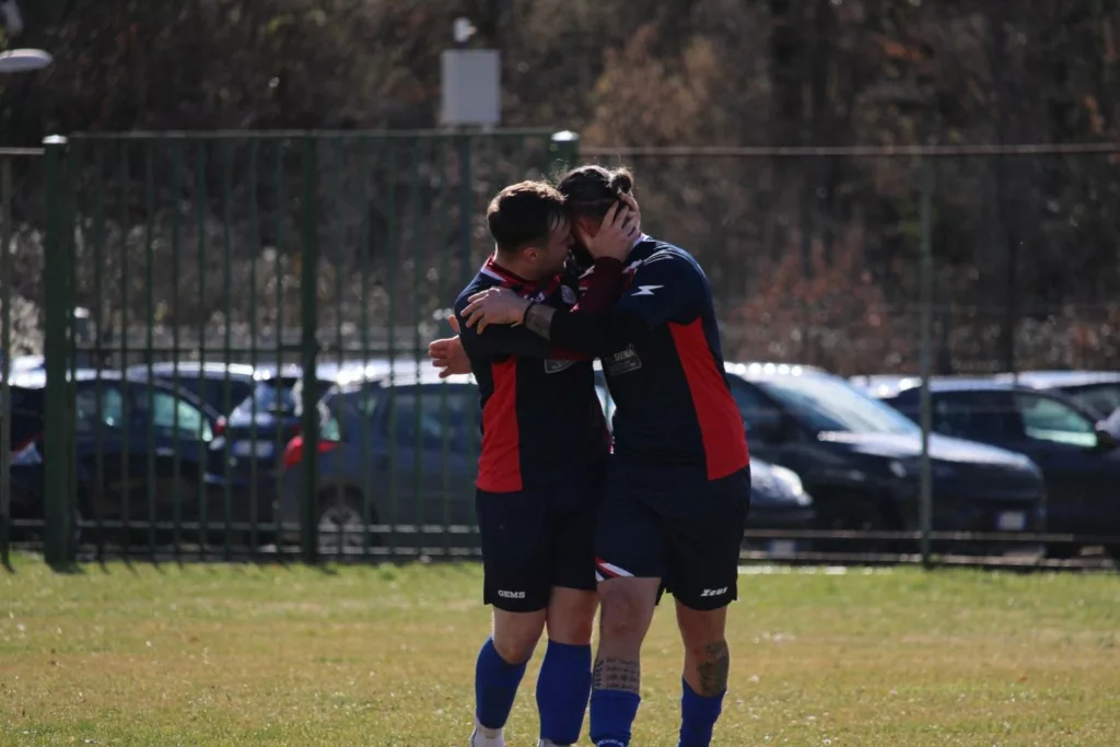 Due giocatori in maglia scura abbracciati su un campo da calcio, auto parcheggiate sullo sfondo. Sofia Gasbarri — ASD Montorio Romano — SPORTSHOTS Magazine