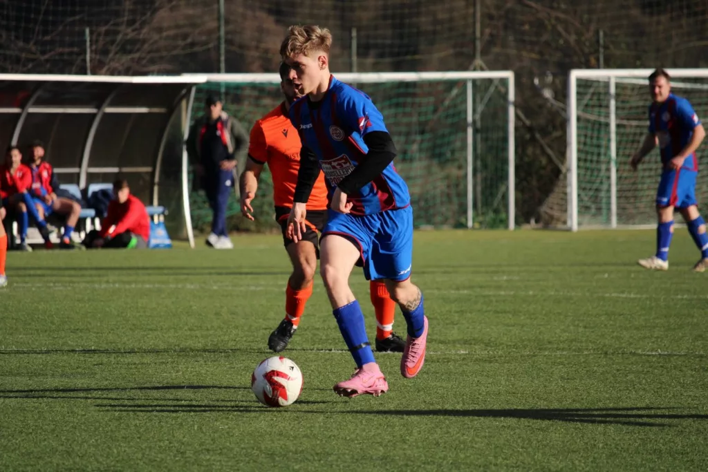 Calciatore in maglia blu e arancione che conduce il pallone, panchina e porta sullo sfondo. Sofia Gasbarri — ASD Montorio Romano — SPORTSHOTS Magazine