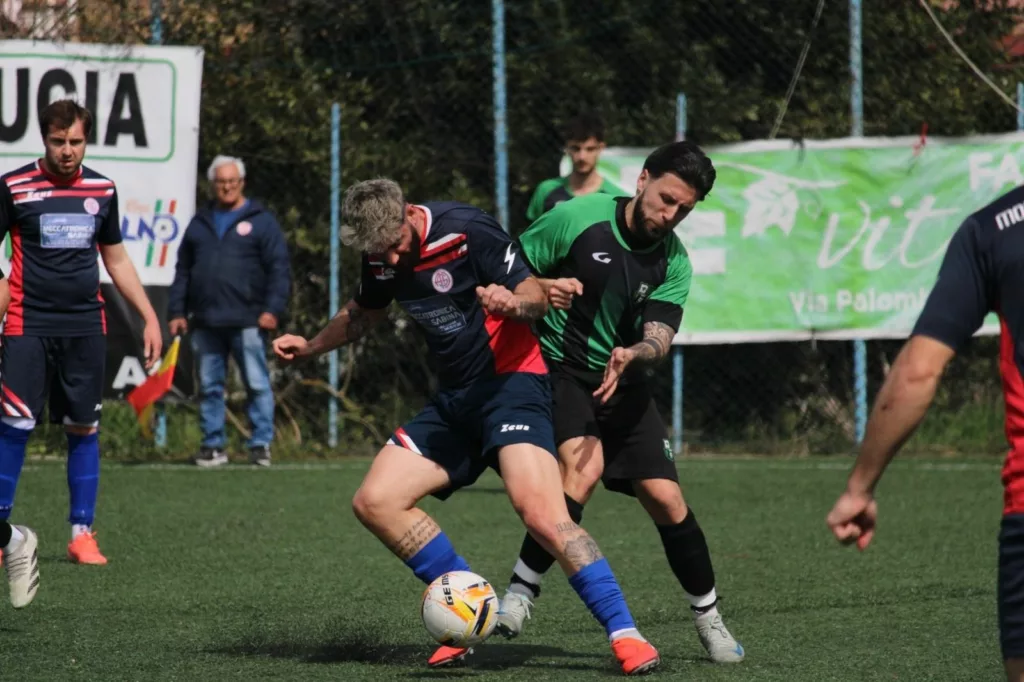 Calciatori in maglia scura e verde che contendono il pallone, un cartello con la scritta in parte visibile sullo sfondo. Sofia Gasbarri — ASD Montorio Romano — SPORTSHOTS Magazine