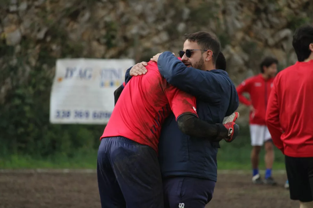 Due uomini abbracciati a bordo campo, uno con giacca rossa e occhiali da sole, altre persone sullo sfondo. Sofia Gasbarri — ASD Montorio Romano — SPORTSHOTS Magazine