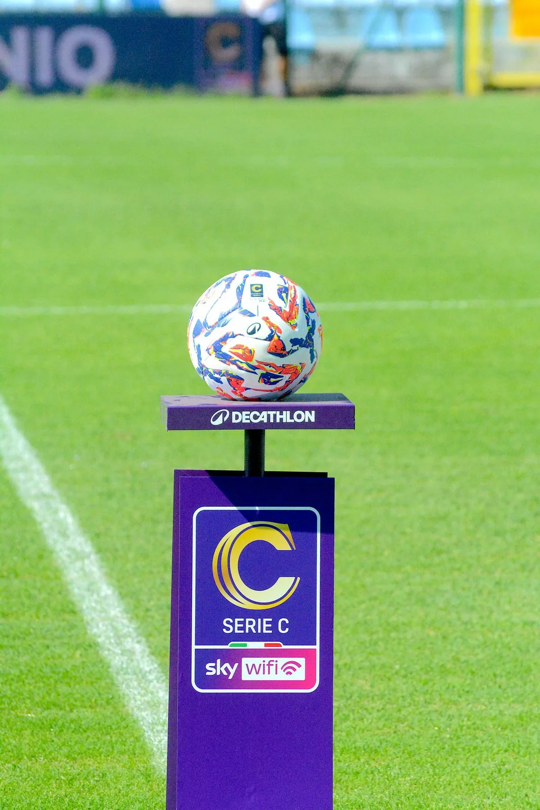 Pallone da gara di Serie C su un piedistallo viola sul prato dello stadio.